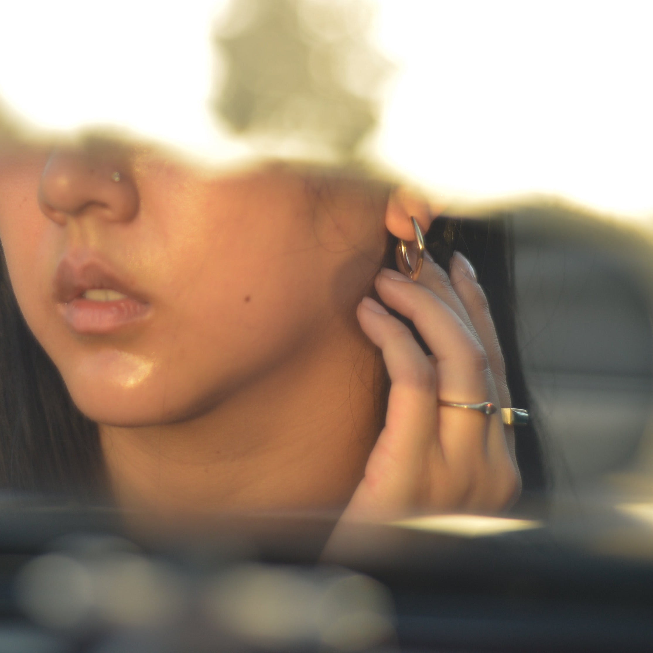 A close-up lifestyle shot of a woman wearing a small gold hoop earring and a gold ring, captured in warm golden-hour light with a soft-focus background.